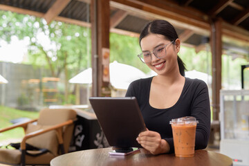 Young beautiful Asian woman working with tablet computer and drinking iced Thai tea in cafe