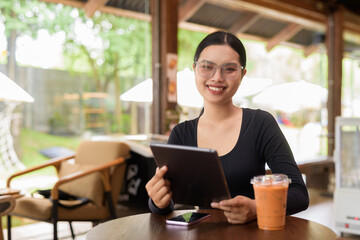 Obraz premium Young beautiful Asian woman working with tablet computer and drinking iced Thai tea in cafe