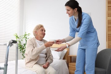 Nurse giving cup of drink to senior woman indoors. Home health care service