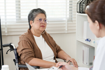 Elderly asian woman sitting in wheelchair listening to female orthopedic doctor during consultation, expressing concern about joint discomfort while receiving supportive medical advice in clinic