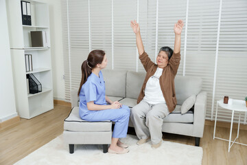 Elderly asian woman stretching both arms upward sitting on sofa during guided exercise by therapist...