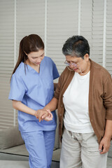 Asian female physiotherapist assisting senior asian female patient with hand support while practicing walking step by step in rehabilitation clinic for muscle recovery and mobility therapy
