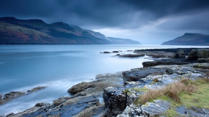 Fototapeta premium Serene coastal scene with calm waters, dark rocky shoreline, and distant mountains under a moody sky