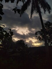 A dramatic sunset or sunrise sky with dark, heavy clouds and a sliver of golden light on the horizon, silhouetting tropical trees.