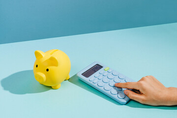 Hand using calculator next to yellow piggy bank on blue background