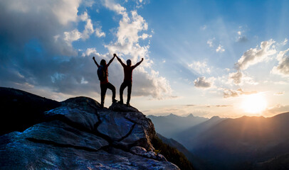 At sunset, two mountain climbers pose on a rock at the summit, raising their arms in joy at their...