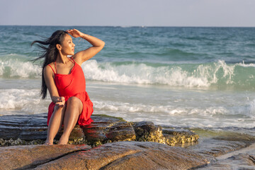 A woman in a red dress sits on a rock on the seashore and looks into the distance