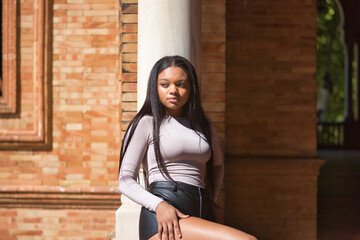 Young, beautiful, African woman, wearing a sweater and leather shorts, looking at infinity, sitting on a railing in the square of Spain in Seville. Fashion concept, beauty, trendy.
