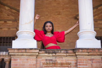 Young, beautiful, African woman in a red dress, looking at camera leaning between two marble...