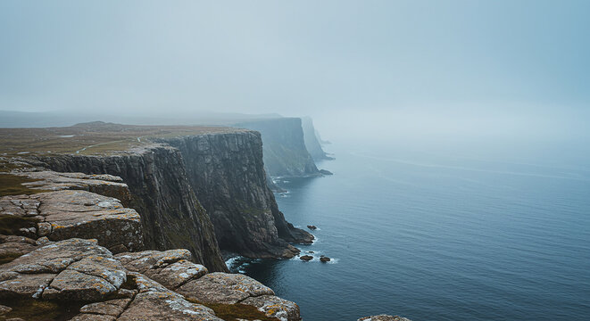 A view of rocky cliffs meeting the ocean under a hazy sky with fog rolling in from the distance