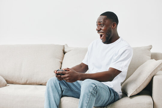 Excited African man playing video games on a couch, showcasing joy and engagement, modern casual outfit, neutral background, perfect for gaming and leisure concepts