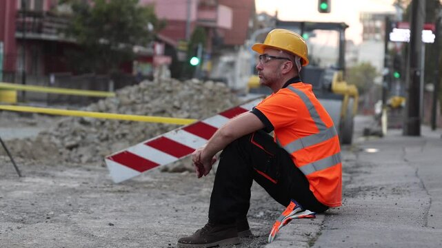 A construction worker in an orange safety shirt and helmet sits on a curb, resting during roadwork in a bustling city.