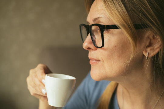 Happy dreamy middle aged woman is staying in living room with cup of black tea or coffee, looking away, Peaceful mature lady enjoying no stress calm positive pastime alone at home.