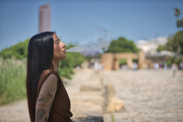 Young woman, brunette, tall, slim and very tattooed, wearing brown dress, vaping, sitting by the river. Concept of youth, beauty, rebelliousness, millennial.