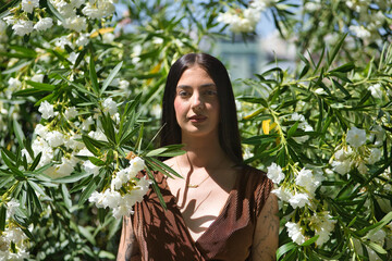 Portrait of young woman, brunette, tall, thin and very tattooed, with brown dress, looking at camera surrounded by white oleanders. Concept of youth, beauty, rebelliousness, millennial.