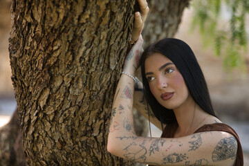 Portrait of a young woman, dark-haired, tall, thin, and heavily tattooed, wearing a brown dress, with a dreamy, sweet, tender, and innocent look, leaning against the trunk of a tree.