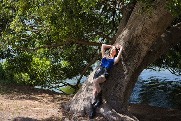 Young woman, brunette, tall, slim, and very tattooed, wearing a blue top, leather shorts, and boots, leaning against the trunk of a large tree soaking up the sun. Concept of youth, beauty, millennial.