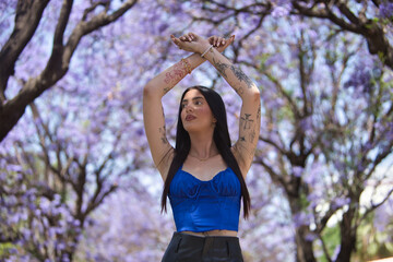 Young woman, brunette, tall, thin and heavily tattooed, wearing a blue top and short leather pants, looking into the distance with her arms crossed above and surrounded by purple bougainvillea trees.