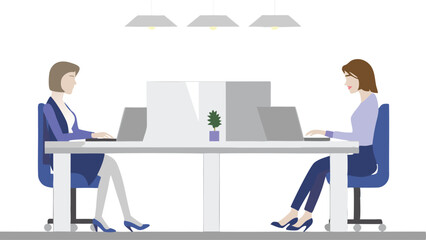 Two women working on laptops at a shared desk in an office with overhead lighting and a small plant