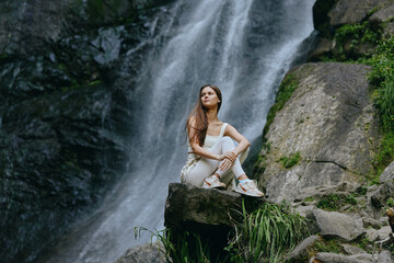 young woman sitting on rock near waterfall in forest, wearing casual white outfit and sneakers,...