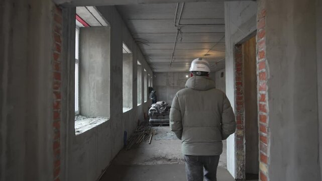 Worker in padded jacket and helmet strolls through unfinished building. Cold concrete space reveals structural stage. Raw construction site