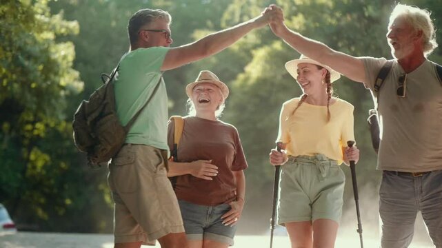 Front view of joyful male and female middle-aged friends giving high-fives and laughing while walking through the forest during a summer hiking adventure.