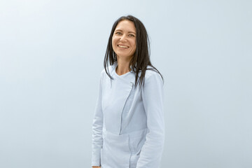 A smiling female massage therapist in a light blue uniform stands against a plain light blue...