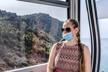 Woman in a face mask and sunglasses riding the Alanya Teleferik cable car with a view of the mountainous landscape, Turkey. The scenic background includes dense greenery and a distant sea