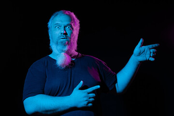 Man with beard pointing to the side with one hand, the other hand on his chest, pink and blue neon lights illuminating him.