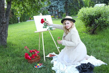 A woman sits on the grass in a garden, painting a red rose on a canvas while surrounded by flowers...