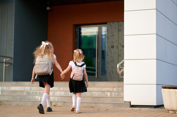 Obraz premium back-to-school girls walking hand-in-hand gleefully towards school entrance wearing backpacks and uniforms. capture of childhood friendship and anticipation of new school year journey