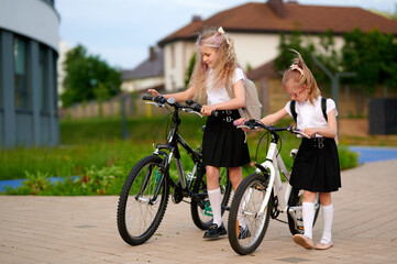 Obraz premium schoolgirls with bicycles outdoors, wearing matching uniforms and backpacks, stand on paved path. girls appear to engage in conversation, suggesting friendship, adventure, active lifestyle.