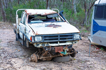 Photograph of an old rusty and destroyed utility vehicle that has been parked and left on a side road adjacent to other discarded items in a large forest area.