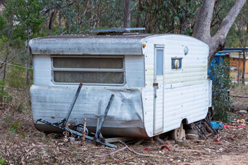 Photograph of an old rusty and destroyed caravan that has been parked and left on a side road adjacent to other discarded items in a large forest area.
