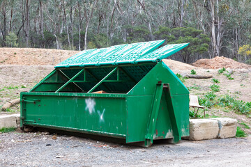 Photograph of a large green steel industrial skip rubbish bin with lids open left on the side of a dirt road in a regional forest. 