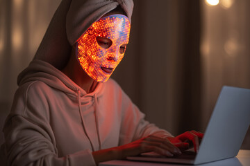 Young woman having red LED light facial therapy treatment. Person wearing LED face mask for beauty and skin rejuvenation. anti ageing collagen at home skincare. Remote working at laptop