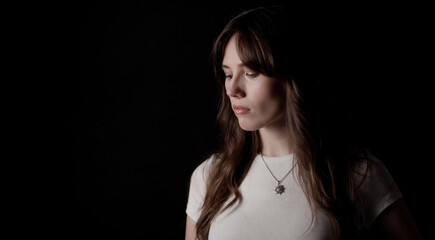 Naklejka premium Studio portrait of a young woman wearing a white t shirt and a necklace with a pendant, looking down against a black background