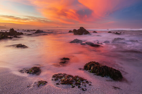 View of a dramatic sunset painting the sky in fiery hues over the rocky coastline, reflecting on the misty ocean waters, Hermanus, Western Cape, South Africa.