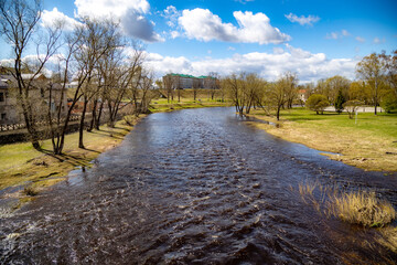 Spring view of the fast-flowing Pskova River after the dam spillway. Foam patterns on water, grassy...