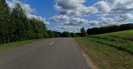 Fototapeta premium Road in the countryside, roadside and asphalt, green forest and fields, blue sky and clouds 