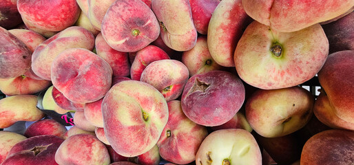 Fresh flat peaches with fuzzy skin in soft pink, red, and creamy tones, stacked closely together in...