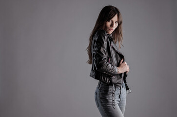 Stylish young woman with long brown hair wearing a black leather jacket and jeans, posing against a...