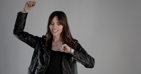 Happy young woman with black leather jacket dancing with eyes closed on gray background