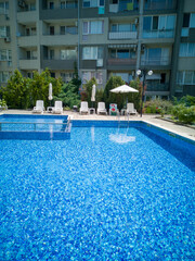 Clean outdoor pool with clear blue water, sun loungers, and umbrellas near modern apartment building. Peaceful summer vibe and relaxation area.