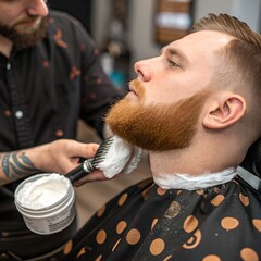 A young Caucasian man with a well-groomed red beard sits in a barber chair. A barber applies shaving cream with a brush. The setting is a modern barbershop.