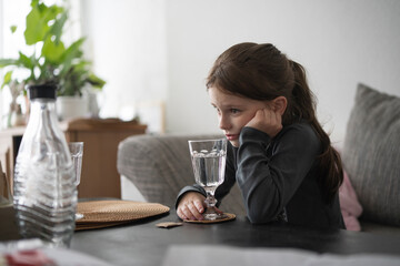 Sad little girl with a ponytail sitting at the table, resting her head on her hand, holding a glass of water. A water carafe stands before her. Concept: childhood sadness