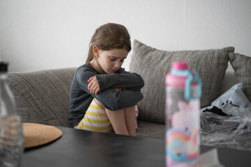 Sad girl with a ponytail sitting on a couch hugging her knees. Concept: childhood vulnerability,...