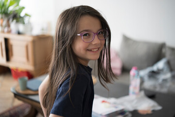 Happy girl wearing glasses in a cluttered bedroom. Concept: natural moments, relaxed atmosphere, childhood spontaneity, genuine happiness.