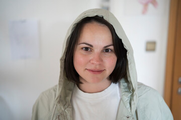 Smiling friendly young woman in a jacket with hood standing near a door indoors. Concept: warmth, approachability, casual comfort, cozy atmosphere, positive energy.