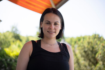 Smiling young woman in tank top and jeans standing in front of an open awning with sky in the background. Concept: summer mood, relaxed living, freedom, home comfort, bright days.